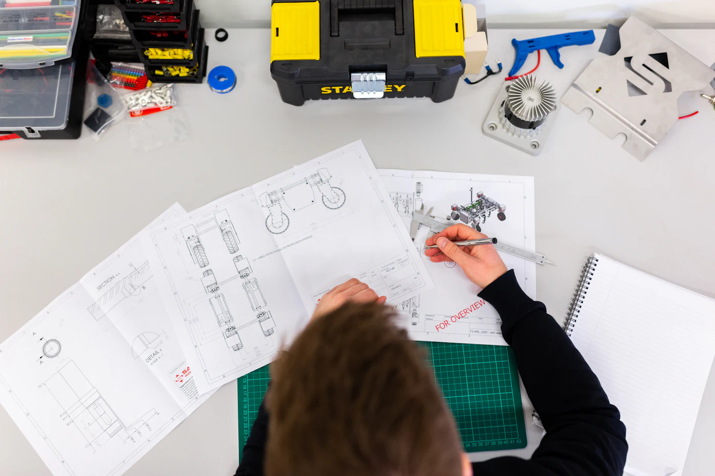An engineer sketching mechanical drawings by hand on a desk covered in technical diagrams.