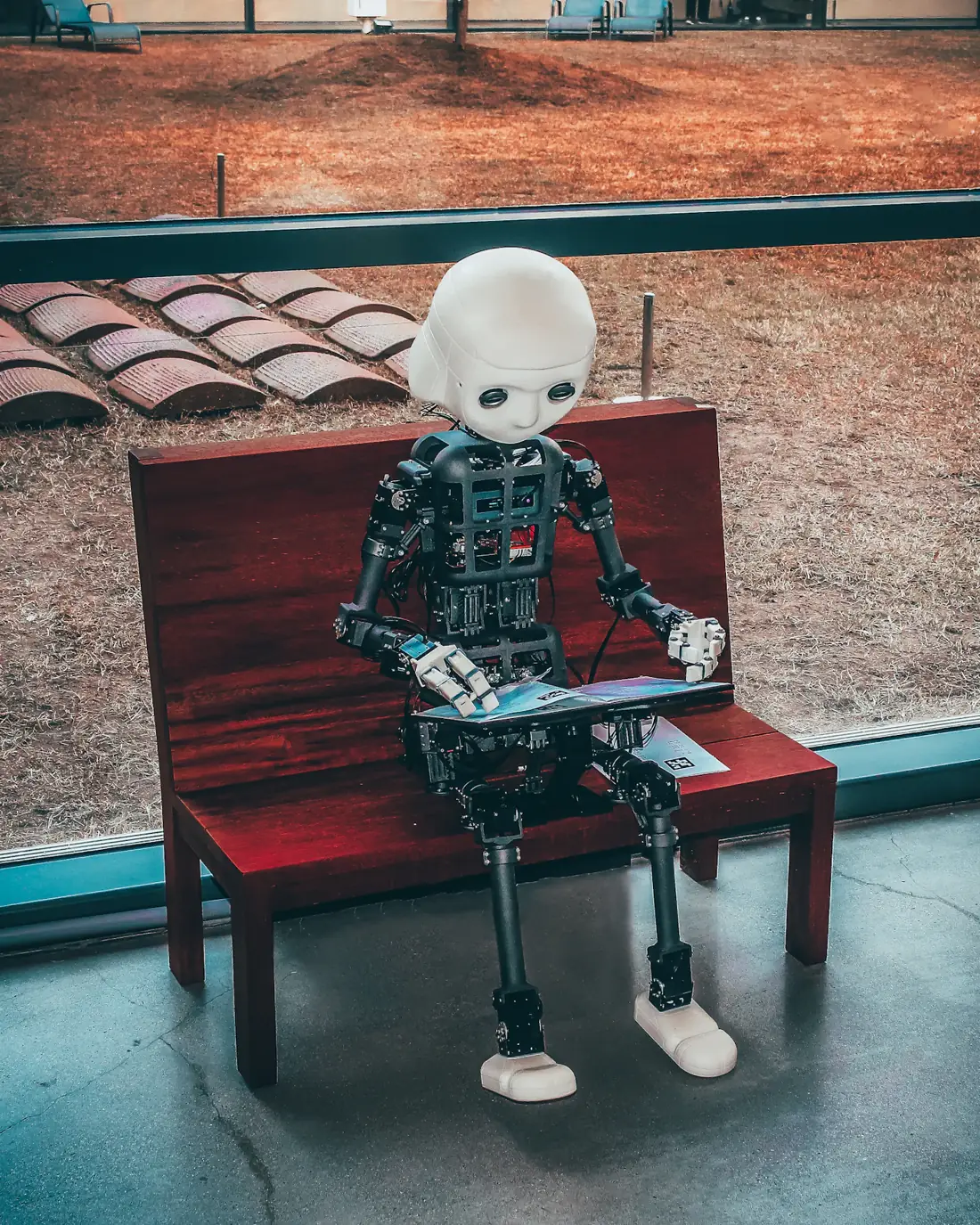 A slender white humanoid robot sitting upright on a red wooden bench outdoors, hands resting on its knees.